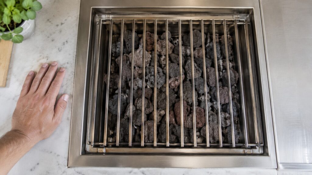 A hand resting beside the lava rocks and the grate of a cooled grill module, seen from above, no text or markings