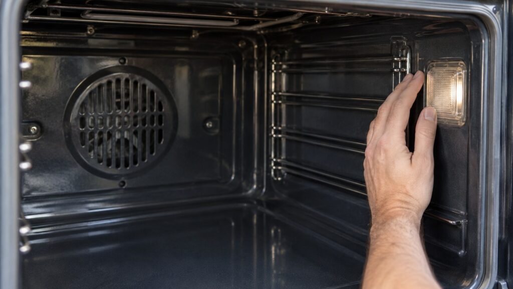 A hand resting beside a small lamp cover on the side wall of a cooled, empty oven, no text or markings